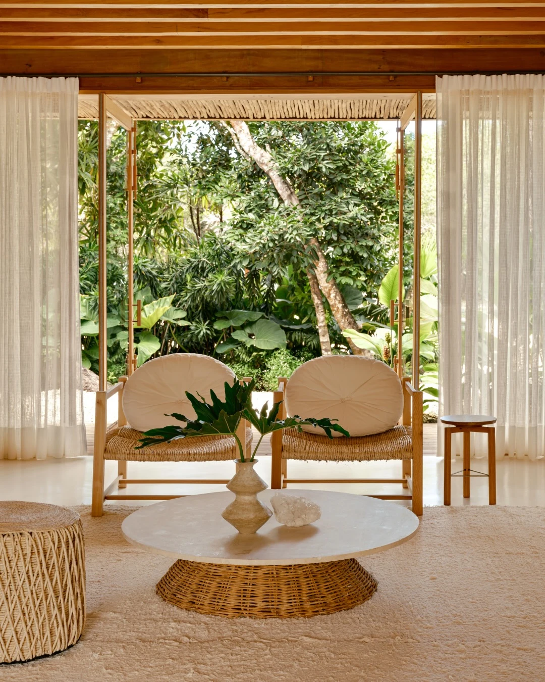 Tropical minimal living room with woven chairs, a low natural-fibre table and open doors framing a lush garden