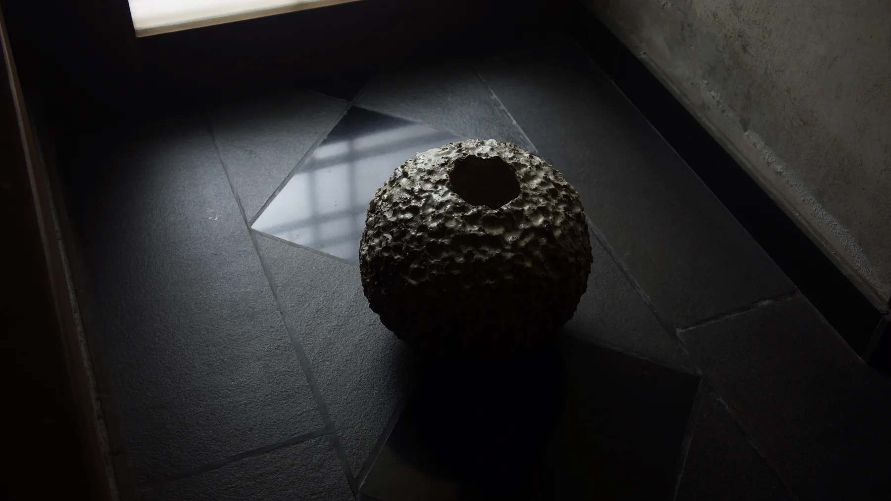 A textured hollow ceramic vessel lit by a window on a dark stone floor